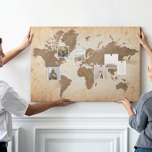 May include: A couple are hanging a world map on a white wall. The map is made of burlap and has a vintage look. There are photos pinned to the map, showing different locations around the world.