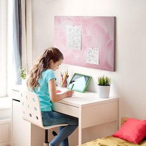 May include: A rectangular pink notice board with a textured, brushstroke-style finish mounted on a white wall. Two white papers with handwritten sketches are pinned to the board surface using small, round, metallic-coloured push pins.