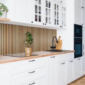 Può includere: Armadietti da cucina bianchi con ante in vetro e ferramenta nera. Una parete a listelli di legno dietro gli armadietti con una piccola pianta in vaso. Un rubinetto e un lavello neri con un tagliere di legno sul bancone.