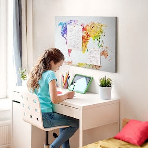 May include: A young person sits at a white desk, writing. A colorful world map art piece hangs on the wall. A tablet, pencils, and a small potted plant are on the desk. The room has a window with a curtain.