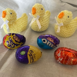 May include: Three yellow knitted chicks with white ribbon bows and five chocolate Easter eggs wrapped in foil. The eggs are Cadbury Creme Egg, Cadbury Oreo, Cadbury Caramel, and Lindor.