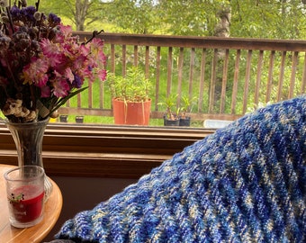 Könnte beinhalten: Eine blaue und weiße gestrickte Wolldecke mit Chevron-Muster, die auf einem Sofa vor einem Fenster mit Blick auf eine Holzterrasse und Bäume liegt.