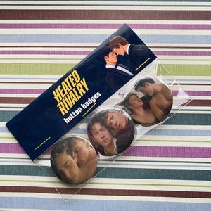 Può includere: Un pacchetto di spille rotonde con immagini di due uomini. La confezione include un'etichetta blu scuro con il testo "HEATED RIVALRY button badges" in giallo e bianco. Le spille hanno una finitura lucida.
