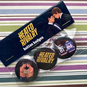 May include: A collection of three round button badges and a rectangular card, all featuring the text "HEATED RIVALRY button badges". The badges display images of two men and the text "HEATED RIVALRY". One badge has a hockey jersey design.