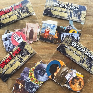 May include: Four clear plastic bags filled with sticker packs. Each bag has a sticker pack with a black and white illustration of a house in the woods with the text "Occult, Creepy & Creepy" on it. The stickers in the bags are various colors and feature illustrations of eyes, hands, faces, and other spooky imagery.