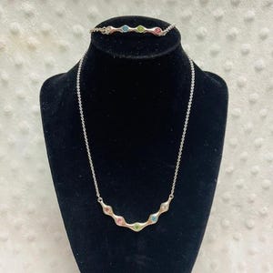 May include: A silver-toned necklace and bracelet set. The necklace features a delicate chain with a curved pendant adorned with colorful gemstones. The matching bracelet has a similar design. The gemstones are pink, green, blue, and clear.