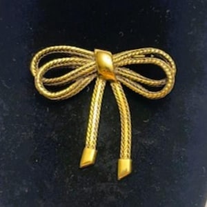 May include: A gold-toned bow brooch with a textured, woven design. The bow features a central rectangular accent and two dangling, rectangular-tipped streamers. The brooch is set against a dark background.