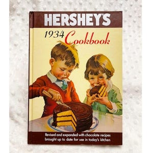 May include: A vintage 1934 Hershey's Cookbook with a cover featuring two children, one cutting a chocolate cake and the other taking a bite. The text on the cover reads "HERSHEY'S 1934 Cookbook" and "Revised and expanded with chocolate recipes brought up to date for use in today's kitchen."