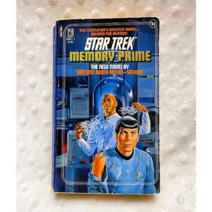 May include: A vintage paperback book titled "Star Trek: Memory Prime." The cover features illustrations of two characters and the text "The Federation's Greatest Minds Marked for Murder!" in blue and yellow.