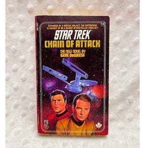 May include: A vintage paperback book titled "Star Trek: Chain of Attack" by Gene DeWeese. The cover features a purple and black space scene with the Star Trek Enterprise and portraits of two characters. The book is a Pocket Books edition.