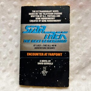May include: A paperback book titled "Star Trek: The Next Generation: Encounter at Farpoint." The cover features the Star Trek logo in blue and silver, with text about the novel's origin and author. The background is black with white stars.