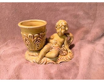 May include: A ceramic figurine of a cherub sitting on a base with a small, decorative pot. The figurine and pot are both a light yellow color.