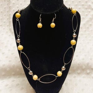 May include: A necklace and earring set displayed on a black mannequin. The necklace features oval silver links, yellow spherical beads, and silver ball beads. The earrings have matching yellow spherical beads. The jewelry set is a neutral color.
