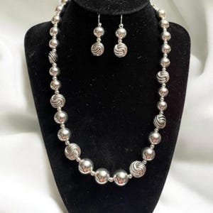 May include: A silver-toned necklace and earring set displayed on a black velvet bust. The necklace features a strand of round beads, interspersed with textured, swirled beads. The matching earrings have a similar design, with a small, swirled bead.