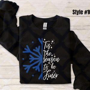 May include: Black sweatshirt with a blue snowflake design and the phrase "Tis the season to be Finer" in white script. The long-sleeved shirt has a crew neck and is a winter-themed apparel item.