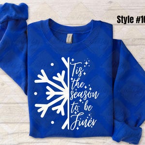 May include: Royal blue sweatshirt with a white snowflake design and the text "Tis the season to be finer." The long-sleeved top has a diamond pattern and rolled cuffs. Style #106 is printed in the upper right corner.