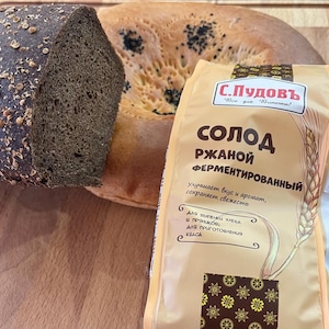 May include: A bag of rye malt fermenting powder with a brown and white label. The label reads "S. Pudov" and "Rye Malt Fermented". The bag is next to a loaf of dark rye bread and a flatbread.
