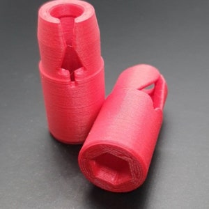 Könnte beinhalten: Zwei rote, 3D-gedruckte Kunststoff-Sechskantmutternhalter. Die Halter haben einen Schlitz oben und eine sechseckige Öffnung unten.