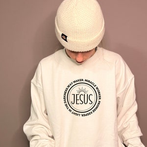 May include: White sweatshirt with a black circle graphic featuring the word "JESUS" in a sunburst design. The circle is surrounded by the text "Light in the darkness, Way Maker, Miracle Worker, Promise Keeper".