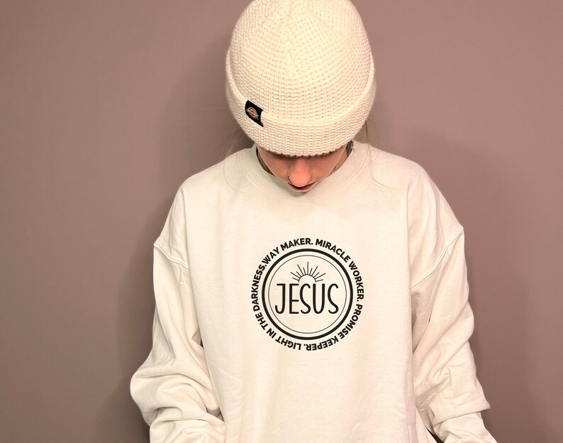 May include: White sweatshirt with a black circle graphic featuring the word "JESUS" in a sunburst design. The circle is surrounded by the text "Light in the darkness, Way Maker, Miracle Worker, Promise Keeper".