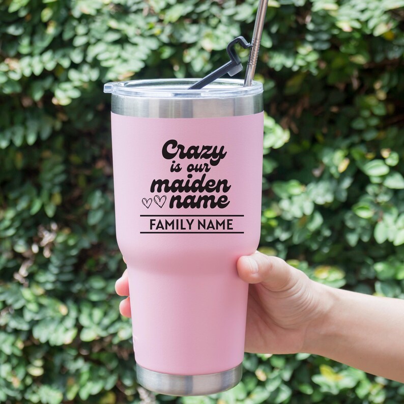 May include: A pink insulated tumbler with a silver rim and lid. The tumbler has the text "Crazy is our maiden name" with two hearts and a line for a family name. A black straw and straw holder are visible.