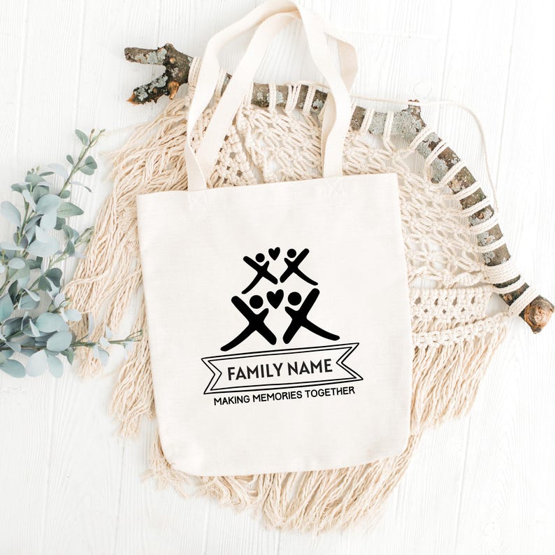 May include: Cream-colored tote bag with black graphic of stick figures and hearts. The text "FAMILY NAME" is above "MAKING MEMORIES TOGETHER" in a banner. The bag has two handles and is on a white surface.