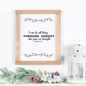 May include: A framed print with a white background and black text that reads "I can do all things THROUGH CHRIST who gives me strength Philippians 4:13". The print is framed in a light brown wood frame and is sitting on a white surface with a white lantern and a green pine branch with white snow.