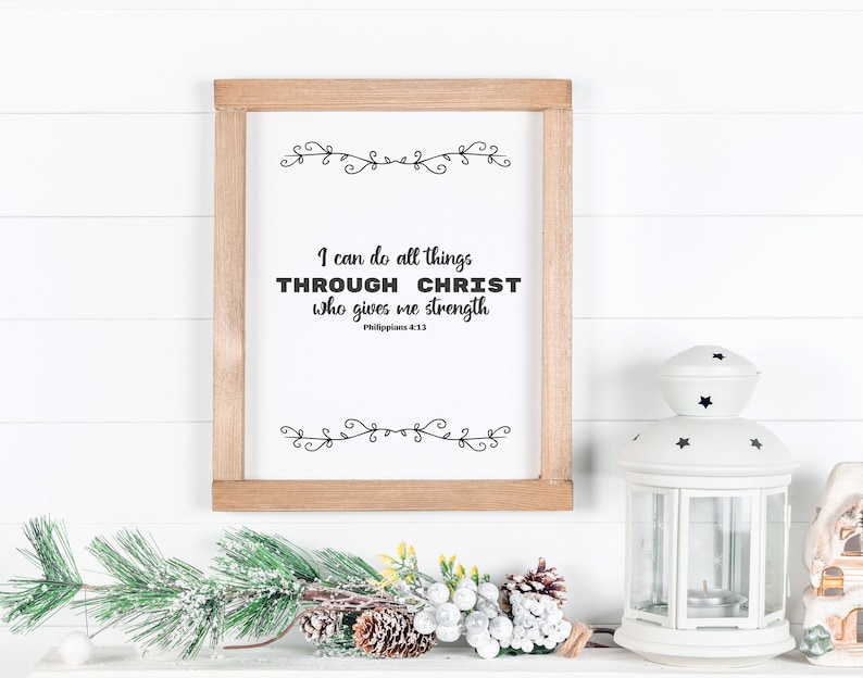 May include: A framed print with a white background and black text that reads "I can do all things THROUGH CHRIST who gives me strength Philippians 4:13". The print is framed in a light brown wood frame and is sitting on a white surface with a white lantern and a green pine branch with white snow.