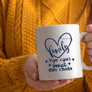 May include: White ceramic mug with a blue heart-shaped design that says "Family". Below the heart, the mug reads "You can't break this chain" in blue script. The mug has a white handle and is held by a person wearing a yellow sweater.