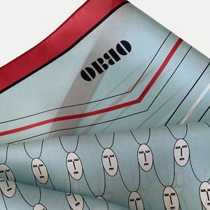 May include: A light blue silk scarf with a red border and the word "OBBO" in black. The scarf features a repeating pattern of white oval faces with black outlines and a geometric design.