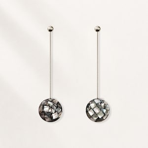 May include: A pair of long drop earrings featuring thin silver-toned metal chains attached to spherical pendants. Each sphere is covered in a mosaic of iridescent, multi-tonal gray and white shell tiles with a smooth, reflective finish.