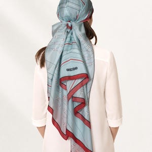 May include: A light blue silk headscarf featuring a repeating pattern of stylized faces, thin horizontal lines, and a bold red border. The scarf is tied at the back and displays the black text ORAO printed clearly on the lower section of the fabric.