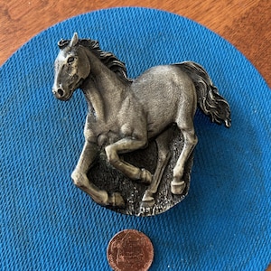 May include: A silver-toned metal belt buckle featuring a detailed sculpture of a running horse. The buckle is approximately the size of a US penny.