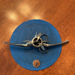 Puede incluir: Una hebilla de cinturón de avión de plata con una hélice y alas. La hebilla está en un círculo azul.