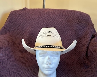 Puede incluir: Un sombrero vaquero blanco con una banda marrón y costuras negras. El sombrero tiene un diseño tejido en la corona.