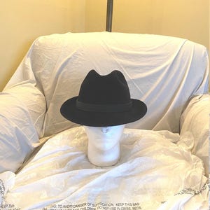 Pode incluir: Um chapéu fedora preto com uma faixa de grosgrain preta. O chapéu está sentado em uma cabeça de manequim branca.