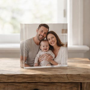 May include: A clear acrylic block displaying a family portrait of a smiling couple and a baby. The photo cube sits on a wooden table, with a window and bed visible in the background.