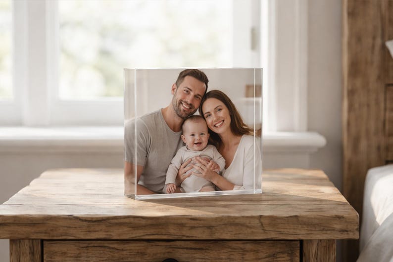 May include: A clear acrylic block displaying a family portrait of a smiling couple and a baby. The photo cube sits on a wooden table, with a window and bed visible in the background.