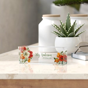 Peut inclure: Un porte-nom de bureau en acrylique transparent avec un motif floral compos&eacute; de fleurs rouges, orange et blanches. Le porte-nom indique "Lily Anderson, HR Director."