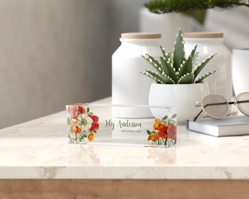 Peut inclure: Un porte-nom de bureau en acrylique transparent avec un motif floral compos&eacute; de fleurs rouges, orange et blanches. Le porte-nom indique "Lily Anderson, HR Director."