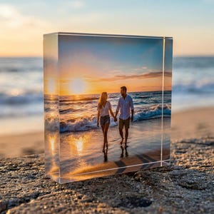 Op de afbeelding: Een helder acrylblok met een foto van een stel dat bij zonsondergang over het strand loopt. De afbeelding staat tegen een achtergrond van een zandstrand, oceaangolven en een levendige oranje en gele lucht.