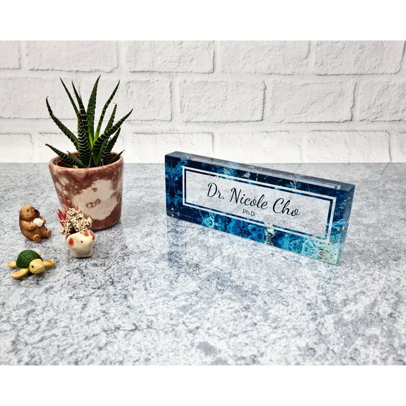 May include: A blue and white acrylic desk nameplate with a white border and the text "Dr. Nicole Cho, Ph.D." in black.