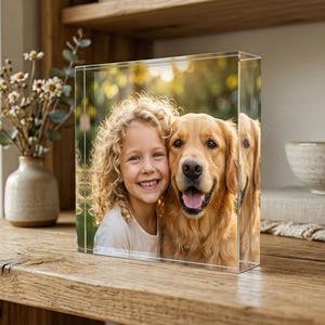May include: A clear acrylic photo block displaying a portrait of a smiling girl and a golden retriever dog. The image is set against a blurred outdoor background. The block is on a wooden surface, with a vase of flowers and a decorative bowl in the background.