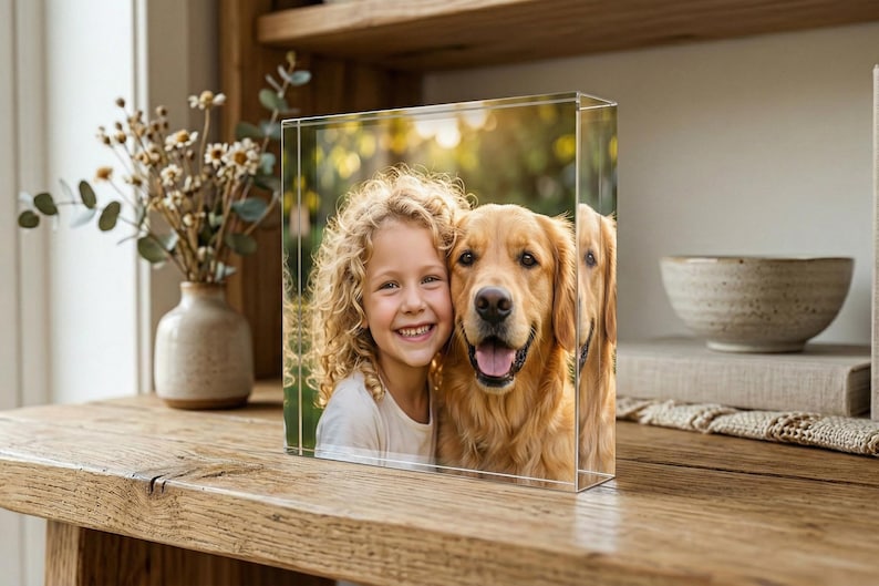 May include: A clear acrylic photo block displaying a portrait of a smiling girl and a golden retriever dog. The image is set against a blurred outdoor background. The block is on a wooden surface, with a vase of flowers and a decorative bowl in the background.
