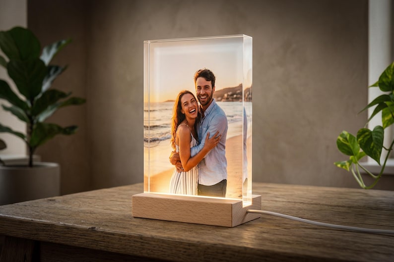 May include: A rectangular photo block with a picture of a couple embracing on a beach at sunset. The photo is encased in clear acrylic and sits on a wooden base with a light. A white cord extends from the base.