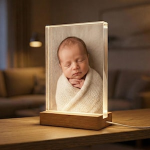 May include: A clear acrylic photo block with a sleeping baby wrapped in a cream-colored blanket. The block is illuminated from the base by a warm light, and sits on a wooden base. The photo block is on a wooden table.