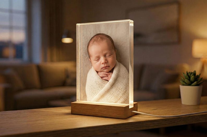 May include: A clear acrylic photo block with a sleeping baby wrapped in a cream-colored blanket. The block is illuminated from the base by a warm light, and sits on a wooden base. The photo block is on a wooden table.
