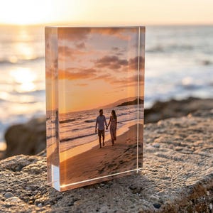 May include: A rectangular acrylic block displays a photograph of a couple walking on a beach at sunset. The image is set against a backdrop of the ocean and a vibrant, orange sky. The block is resting on a rough, stone surface.