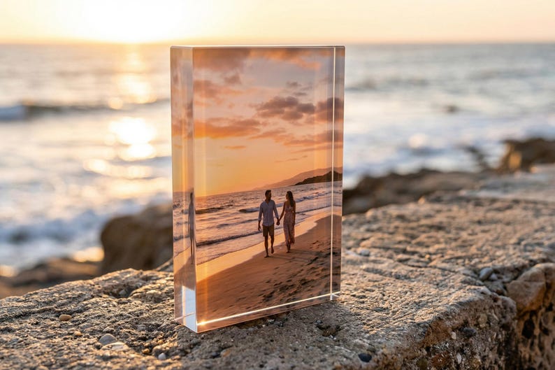 May include: A rectangular acrylic block displays a photograph of a couple walking on a beach at sunset. The image is set against a backdrop of the ocean and a vibrant, orange sky. The block is resting on a rough, stone surface.