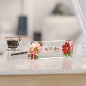 May include: A clear acrylic desk nameplate with a floral design featuring red and white flowers. The nameplate reads "Rachel Adams, Surgeon".